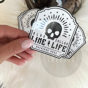 Pu&ograve; includere: Un adesivo bianco con un teschio nero e il testo "Blue Collar Life American Made" in un font in stile vintage.
