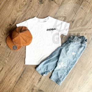 Peut inclure: Un t-shirt blanc avec le texte "And Little Souls" en noir, associ&eacute; &agrave; un jean bleu clair d&eacute;chir&eacute; et une casquette de baseball en velours c&ocirc;tel&eacute; marron. La casquette a un &eacute;cusson circulaire.