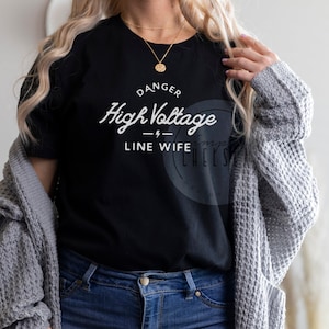May include: A black t-shirt with white text that reads "Danger High Voltage - Line Wife". The person wearing the shirt is also wearing blue jeans and a gray cardigan.