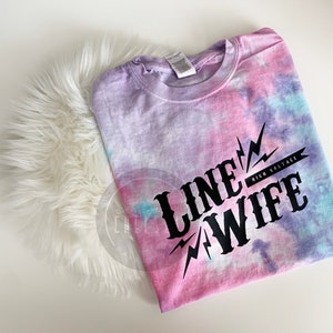 May include: A tie-dye t-shirt in shades of pink, purple, and blue with the text "LINE WIFE HIGH VOLTAGE" printed in black.