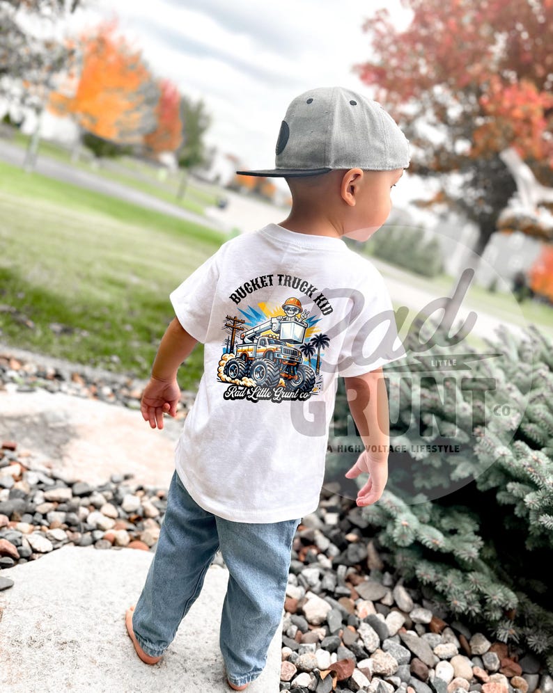 Peut inclure: T-shirt blanc avec un graphisme color&eacute; d'un camion nacelle et le texte "Bucket Truck Kid". Le t-shirt est port&eacute; par un enfant portant un jean bleu et une casquette de baseball grise. Le t-shirt porte &eacute;galement le texte "Rad Little Grunt co."