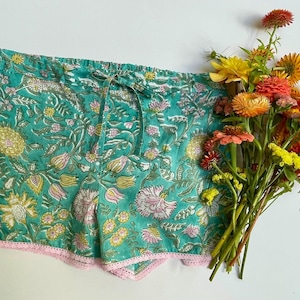 May include: Teal shorts featuring a multi-coloured floral print in shades of pink, yellow, and green. The shorts have an elastic waistband with a matching fabric drawstring and a delicate light pink lace trim along the bottom hem edges.
