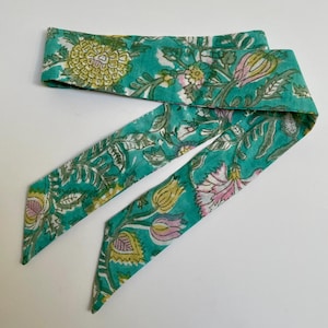 May include: A long, narrow fabric hair ribbon featuring a vibrant teal background with a repeating floral pattern in shades of yellow, pink, and white. The accessory has pointed ends and displays a detailed botanical print with leaves and blossoms.