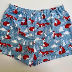May include: Light blue boxer shorts featuring an all-over print of red foxes, white rabbits, and white pine trees. The garment has an elasticated waistband and a relaxed fit, showcasing a repeating pattern of woodland animals and snowy clouds.