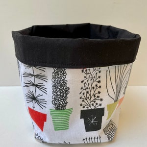 May include: A black and white fabric storage basket with a black fabric lining. The basket features a pattern of potted plants with different coloured pots.