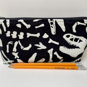 Peut inclure: Trousse noire avec un imprimé de squelette de dinosaure blanc. La pochette zippée présente un motif répété d'os et un crâne de dinosaure. Deux crayons orange avec le texte "STAEDTLER 173 HB" sont devant la trousse.