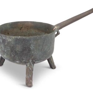 May include: A weathered, antique metal cooking pot with three short legs and a long, flat handle. The pot has a rounded body and a slightly uneven rim, suggesting it is handmade. The metal has a dark, aged patina.