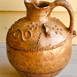 May include: A tan-colored, antique ceramic pitcher with a handle and spout. The pitcher features decorative embossed patterns, including dots and swirls, and a textured surface. The jug has a crack running down the front.