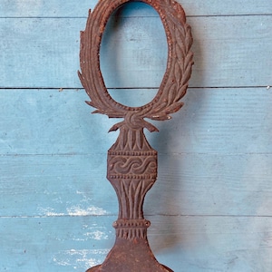May include: A rusty metal decorative piece with an oval frame and a base. The frame is decorated with a leafy design and the base has a wave pattern.