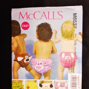 May include: McCall's sewing pattern M6537 for baby diaper covers. The pattern features three babies wearing animal-themed diaper covers: a dog, a pig, and a bear. Additional diaper cover designs include a duck, a dog, and a cat. The pattern is labeled "EASY".