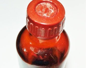 May include: A small, brown glass bottle with a red cap. The bottle is filled with a dark liquid. The cap is round and has ridges around the edge. The bottle is sitting on a white surface.