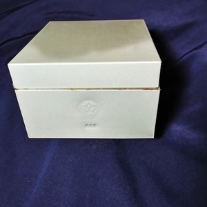 May include: A rectangular, light gray box with a lid. The box has a simple design with a logo and the number "663" embossed on the front. The box is sitting on a dark blue surface.