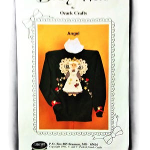 May include: A black sweatshirt with an angel design, framed within a decorative border. The design includes a white angel figure, gold stars, and red accents. The text "Angel" is above the sweatshirt.