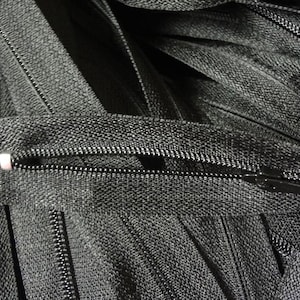 May include: Close-up of several black zippers, some open and some closed. The zippers are made of a dark gray fabric with a metal zipper pull. The image shows the details of the zipper teeth and the fabric texture.