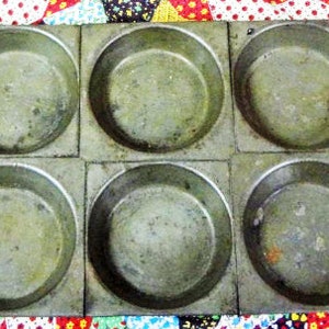 May include: A vintage, six-cup muffin tin made of aged metal. Each cup is round and set within a square frame. The metal has a weathered, greenish-gray patina, suggesting age and use. The tin is designed for baking muffins or cupcakes.