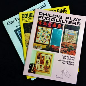 May include: Three books on a black surface. The book titles are "One Patch Beyond", "Double Designs", and "Child's Play for Quilters". The cover of "Child's Play for Quilters" has images of quilts and the text "An Idea Book for Quilters, A Learning Book for Children".