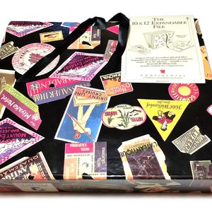 May include: A black expandable file folder decorated with colorful travel-themed stickers. The file has a black ribbon handle and a white label that reads "The 10 x 12 Expandable File". The stickers feature various destinations.