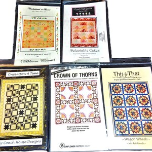 May include: A collection of quilt pattern packets, each sealed in a plastic sleeve. The packets feature various quilt designs, including "Checkerboard in Bloom," "Delectable Cakes," and "Wagon Wheels." The packets display colorful quilt images and text.