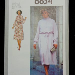 May include: A Simplicity sewing pattern envelope, featuring a woman in a light pink dress with long sleeves and a matching belt. The envelope also shows a floral dress design. The pattern number is 8834.