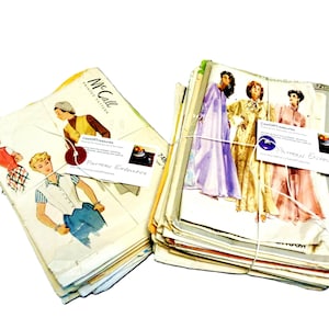 May include: Two stacks of vintage paper sewing pattern envelopes featuring illustrations of clothing designs. The left stack is labeled McCall Printed Pattern. Each stack is tied with white string and includes a white business card with text.