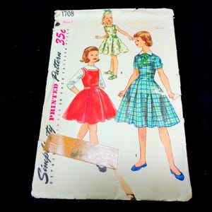 May include: Vintage Simplicity pattern 1708 featuring illustrations of three dresses. The dresses are a red jumper, a green floral dress, and a blue and green plaid dress. The pattern is printed on a white paper with the text "Printed Pattern" and "Simplicity Detail".