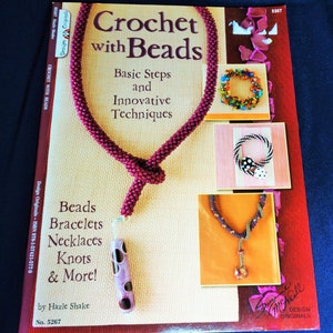 May include: A book titled "Crochet with Beads" featuring a burgundy beaded necklace with a pendant. The cover displays examples of beaded jewelry, including bracelets and necklaces. The book is by Hazle Shake.