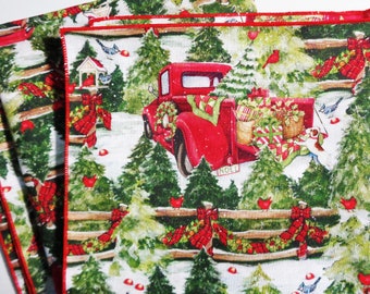 May include: Christmas-themed fabric with a red truck carrying gifts and a wreath. The design features a snowy landscape with evergreen trees, red cardinals, and a wooden fence. The fabric has a red border.