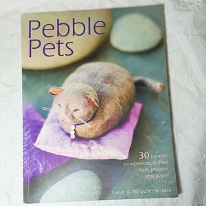 May include: A book cover titled "Pebble Pets" with a purple title. The cover features a crafted pebble cat on a purple pillow. The text below reads "30 lovable companions crafted from pebbles and paper".