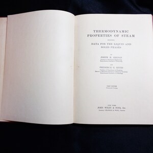 May include: Open book titled "Thermodynamic Properties of Steam" with text "Data for the Liquid and Solid Phases." The book is open to the title page, with text and author names. The book has a red spine and is on a dark blue surface.