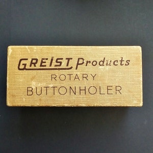 May include: A rectangular box with the text "GREIST Products ROTARY BUTTONHOLER" in brown lettering. The box has a textured, tan surface with a woven pattern. The box is rectangular and appears to be vintage.
