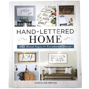 May include: A book titled "HAND-LETTERED HOME" with the subtitle "DIY Wood Signs for Farmhouse Decor." The cover features images of hand-lettered signs with phrases like "gather," "I love you to the moon and back," and "home."