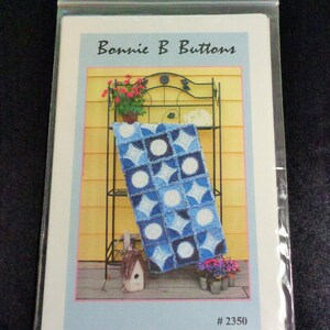 May include: A package featuring a pattern for a blue and white quilt. The package is in a clear plastic bag. The image on the package shows a blue and white quilt draped over a black metal plant stand. The text "Bonnie B Buttons" is at the top.
