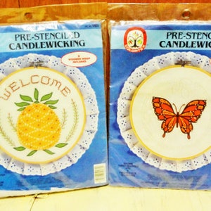 May include: Two pre-stenciled candlewicking kits. One features a pineapple with the word "WELCOME", the other a butterfly. Both are in a wooden hoop with a white lace doily backing. The packaging is blue.