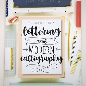 May include: A beginner's guide to lettering and modern calligraphy. The book is surrounded by gold pens, washi tape, and other stationery items. The book has a white cover with black lettering and a banner that says "and".