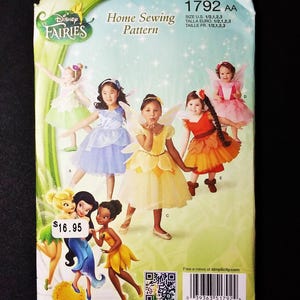 May include: A Simplicity home sewing pattern featuring Disney Fairies. The pattern includes illustrations of children in fairy costumes. The package has the text "Disney Fairies" and "Home Sewing Pattern". The price tag shows $16.95.