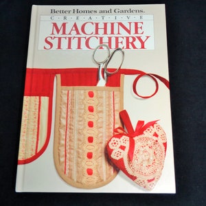 May include: A book titled "Machine Stitchery" by Better Homes and Gardens. The cover features a red and beige sewing-themed design with scissors, a needle holder, and a heart-shaped pincushion.