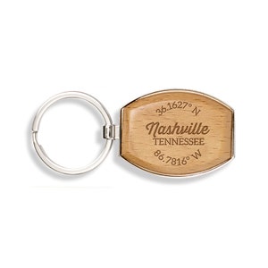 May include: A wooden keychain with a silver-colored ring. The keychain has a light brown, oval-shaped tag with the word "Nashville" in a cursive font, along with the coordinates and "Tennessee" printed below.
