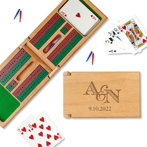 May include: A wooden cribbage board game set with a personalized wooden box. The box has the initials "A&N" and the date "9.10.2022" engraved on it. The cribbage board is open and shows the playing surface with pegs and cards.