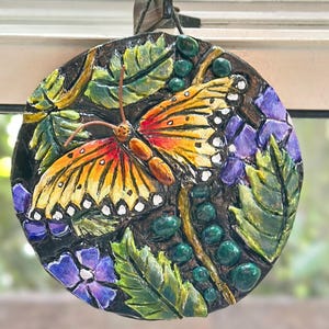 May include: A round, hand-painted decorative plaque featuring a butterfly with orange and yellow wings, detailed with white spots. The butterfly is surrounded by green leaves, purple flowers, and green beads, all set against a dark background.
