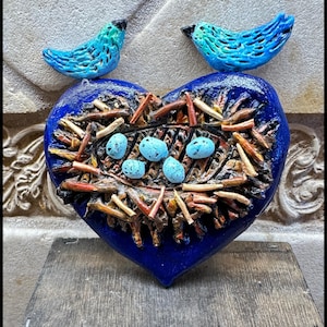 May include: A blue heart-shaped sculpture with a nest design, speckled blue eggs, and two turquoise birds. The nest is made of brown and orange twigs. The birds are perched above the heart, adding a whimsical touch to this decorative piece.