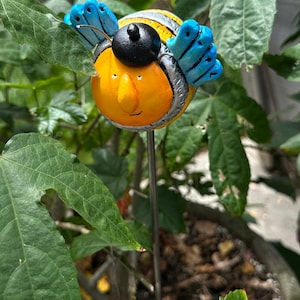 May include: A whimsical garden stake featuring a smiling bee with a yellow body, black head, and blue wings. The bee has a cheerful expression and is mounted on a silver metal stake. The bee is surrounded by green leaves.