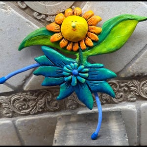 May include: A whimsical, handmade floral sculpture featuring a smiling yellow sunflower with orange petals and a blue and green flower below. The sculpture has green leaves and blue stems, with a textured appearance.