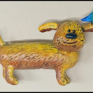 May include: A whimsical, handcrafted sculpture of a dog with a small blue bird perched on its ear. The dog is primarily yellow and brown, with a smiling face and a dark blue nose. The bird is blue and green.