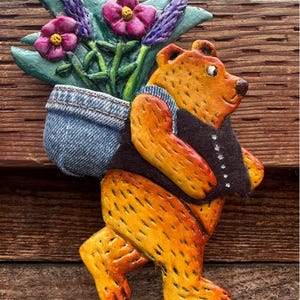 May include: A hand-painted ceramic bear figurine carrying a pot of purple flowers in a blue denim pocket. The bear is walking and has a black vest.