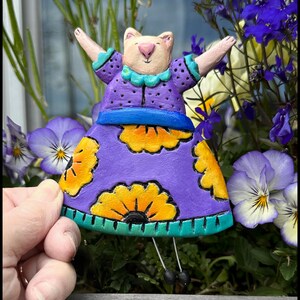 May include: A purple ceramic figurine of a cat wearing a purple dress with yellow flowers and a blue sash. The cat has pink arms and a white face with a pink nose.