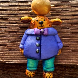 May include: A whimsical figurine of a brown bear wearing a purple jacket with buttons, a purple bow tie, and teal pants. The bear has a golden sun on its head.