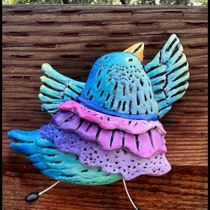 May include: A blue and purple bird figurine with a ruffled skirt, painted with a whimsical design. The bird is perched on a wooden surface.