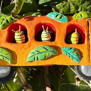 May include: A whimsical, orange school bus figurine with green leaf accents and five bee passengers. The bus has gray wheels and is set against a backdrop of green foliage. The bees are yellow and black.