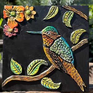 May include: A colorful, 3D hummingbird sculpture with green, orange, and brown accents perched on a branch with green leaves. The sculpture is set against a black background.