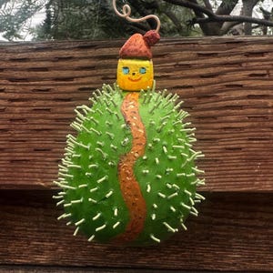 May include: A whimsical, handmade ornament featuring a green, spiky body with a painted orange path down the center. It has a yellow head with blue eyes and a smiling face, topped with a small red hat. A curly wire extends from the top.